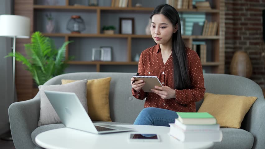 Asian female student listens to online training by video call using laptop taking notes in notebook while sitting on sofa at home. Young woman watches remote learning seminar, lecture, class or course