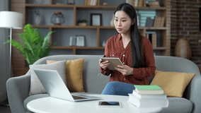 Asian female student listens to online training by video call using laptop taking notes in notebook while sitting on sofa at home. Young woman watches remote learning seminar, lecture, class or course - Powered by Shutterstock - Get 15% off with code: PIKWIZARD15