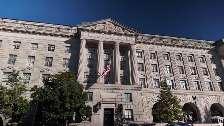 US Department of Commerce building with columns, intricate carvings, and an American flag flying in front. The building has multiple windows, an arched entrance, and is framed by trees.