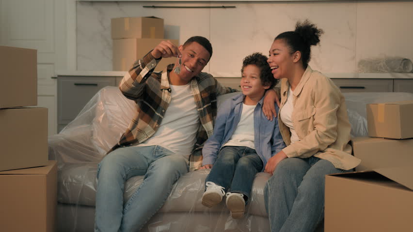 Happy family African American parents little son boy together on couch moving day relocation father holding keys of new own house buy rent apartment surprise mother child kid rejoice credit mortgage
