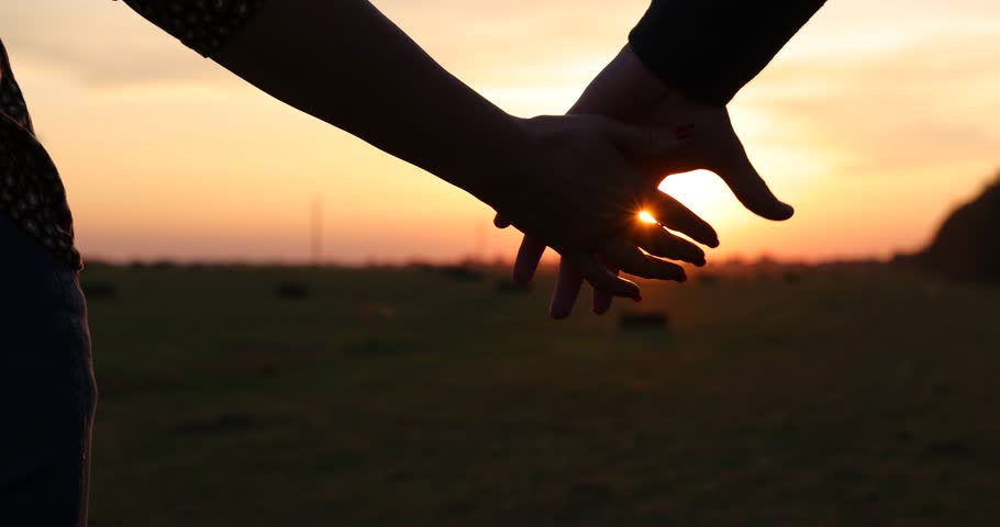 Close-up of the silhouettes of a couple in love, man and woman holding their hands together and the golden sun gently setting below the horizon, romantic scene at sunset, slow motion