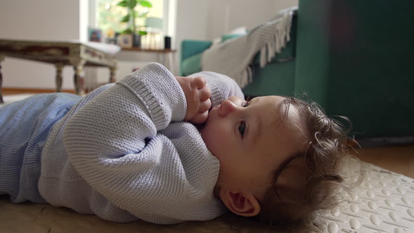Baby lying on floor with hands together, looking up with curious expression, calm indoor family time, baby resting and observing surroundings, playful child in home setting