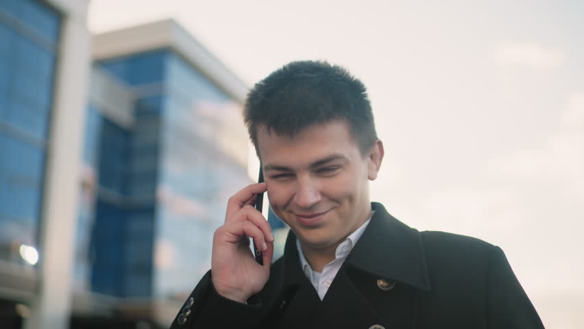 Man in black coat happily talking on phone with excitement while making hand gesture in vibrant urban area with modern glass buildings, soft sunlight, and cheerful atmosphere