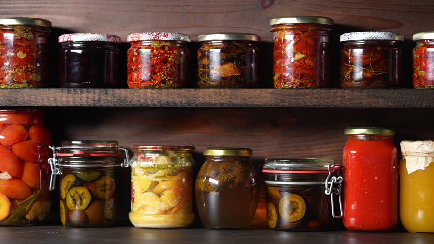 Many canned food in glass jars on wooden shelves in the cellar for winter consumption, close up. Food preservation. Colorful pickled and fermented jars of vegetables, honey, jam and other products