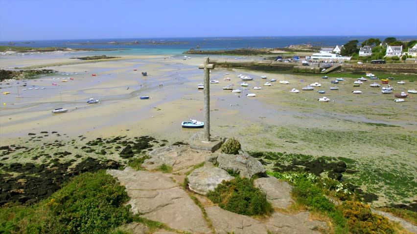 A stone cross stands on a rocky hill overlooking boats stranded on the sand at low tide along the Brittany coast. The drone view captures the expansive bay, shoreline, and nearby village.