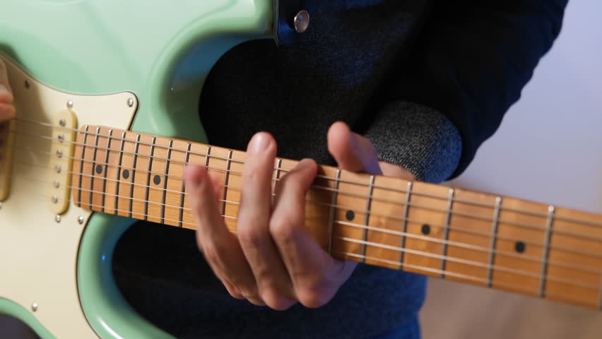 Close-up of male musician soloing on a mint electric guitar at indoor studio, slow motion. Songwriting and practicing music