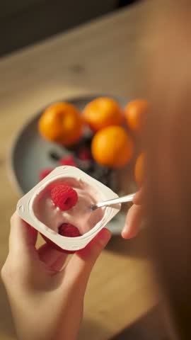 Vertical video: A child eats yogurt with a fresh berry taking a spoon of a glass. The girl uses a spoon in the back to eat fresh solar yogurt from a glass holding in her hand. Food for children