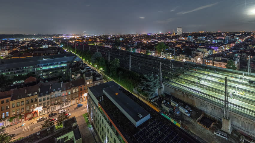 Aerial timelapse of Brussels North station railway tracks with trains arriving and departing. Schaerbeek cityscape panorama at night with illuminated buildings and landmarks. Rising moon. Belgium