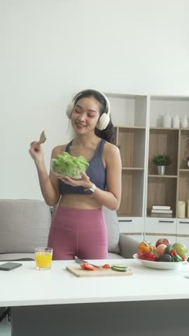 A young Asian housewife enjoys a healthy lifestyle at home, balancing fitness routines with relaxation. She sits on a modern sofa, eating a fresh salad and fruit while embracing healthy habits.
