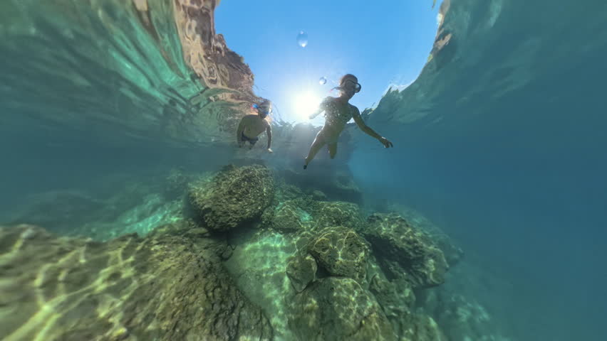 Two snorkelers swim underwater, exploring a rocky seabed illuminated by sunlight. The scene is captured in a wide-angle view, emphasizing the clarity of the water and the beauty of the underwater worl