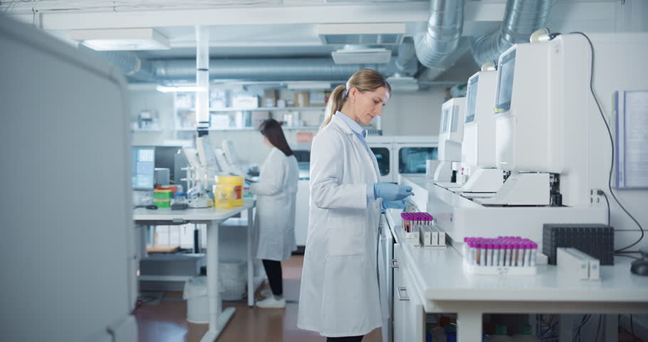 Biomedical Researchers Conduct Experiments in a Modern Laboratory. Female Scientist Preparing a Set of Samples in Test Tubes with Purple Caps. Colleague Analyzes Data on a Computer in the Background