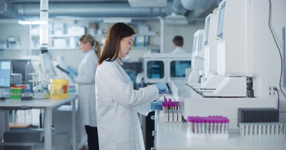 In a Bright and Sterile Hospital Lab, a Young Asian Scientist Carefully Prepares Test Vials for Genetic Analysis as Her Male and Female Colleagues Prepare Medical Equipment in the Background