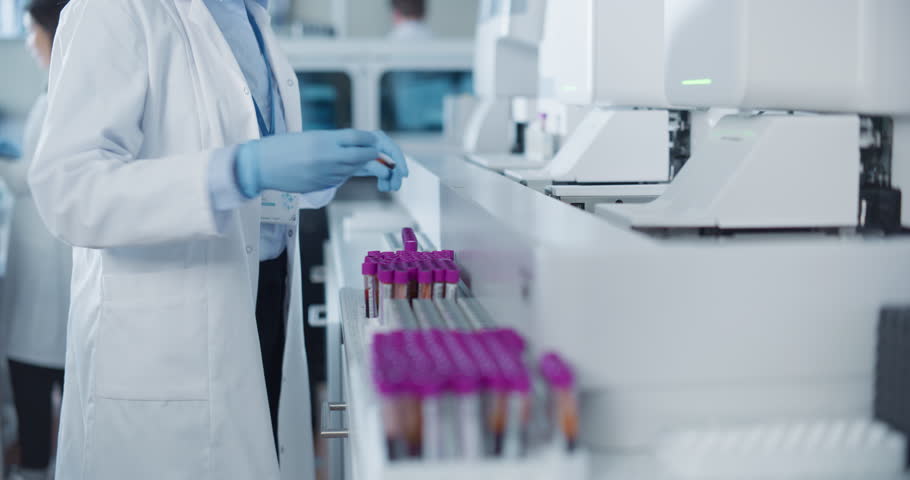 Medical Laboratory Technician in White Coat and Blue Gloves Handles Test Tubes with Purple Caps. Anonymous Worker Feeding Samples Into an Advanced Test Equipment for Scientific Analysis and Research