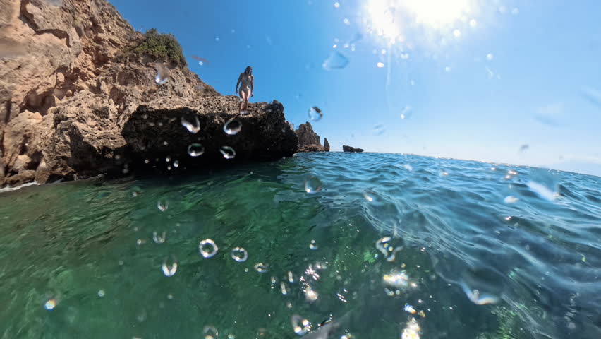 A woman captured mid-jump as she dives from a cliff into the water, with bubbles forming around her as she descends and rocks visible below.