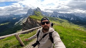 Photographer with backpack and camera taking a selfie with outstretched arm with the odle di eores mountain group in the background - Powered by Shutterstock - Get 15% off with code: PIKWIZARD15
