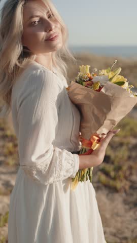 Happy woman in a white dress smiling and holding a beautiful bouquet of flowers on a sunny day at the beach. Perfect for themes of love, happiness, and summer.