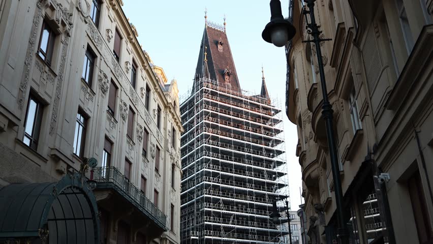 A historic Gothic tower under restoration, surrounded by ornate classic European buildings. The scaffolding highlights ongoing preservation efforts in a charming old-world urban setting.