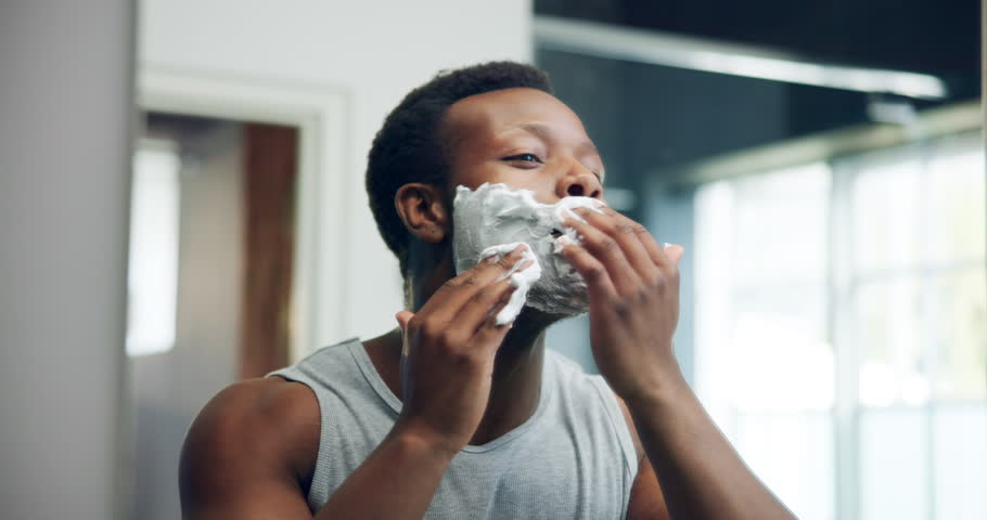 Man, mirror and apply shaving cream on face for hygiene, grooming and morning routine in bathroom. African person, facial cosmetic and foam for hair removal, beauty reflection and skincare at home