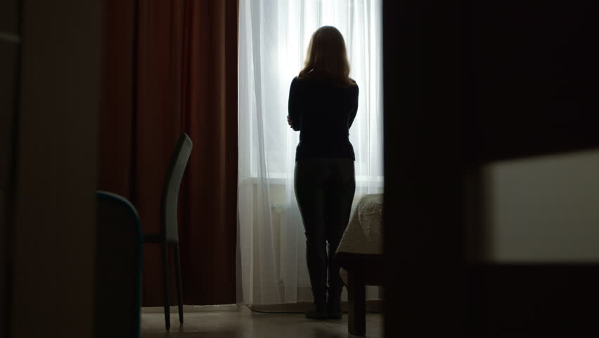 Sad and lonely woman standing at the window in the room. Concept of depression, problems and mental health. Female silhouette.