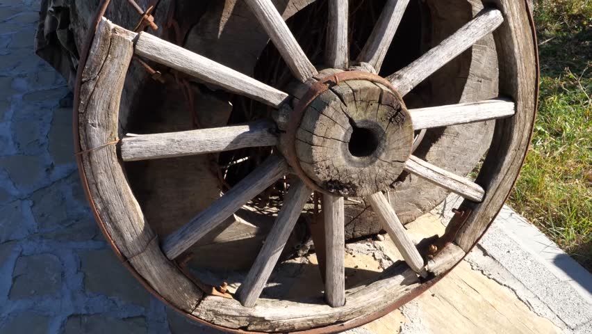Old wooden cart wheel resting on a pathway in the countryside. A rustic wooden cart wheel decoration stands on a stone path surrounded by grass.