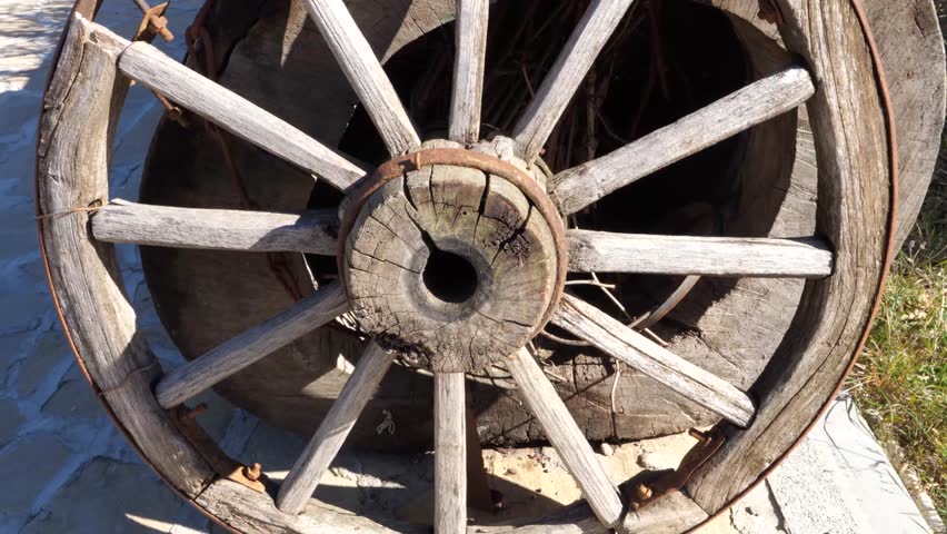 Old wooden cart wheel resting on a pathway in the countryside. A rustic wooden cart wheel decoration stands on a stone path surrounded by grass.
