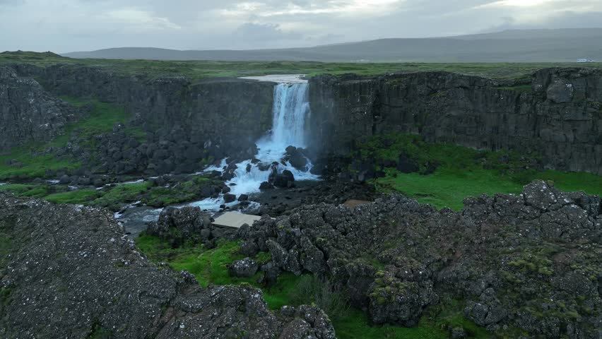 A stunning view of a waterfall in Thingvellir National Park, Iceland, surrounded by rocky cliffs, green fields, and dramatic skies.