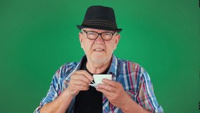 An elderly, 60 year old man, in a hat, holds a cup of coffee in his hands, drinks, smiles.. Slow motion. Green background. - Powered by Shutterstock - Get 15% off with code: PIKWIZARD15