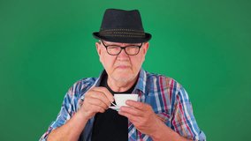An elderly, 60 year old man, in a hat, holds a cup of coffee in his hands, drinks, smiles.. Slow motion. Green background. - Powered by Shutterstock - Get 15% off with code: PIKWIZARD15