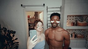 Young multi-ethnic couple with under-eye patches looking into camera and smiling as taking selfies with mobile phone in bathroom at home - Powered by Shutterstock - Get 15% off with code: PIKWIZARD15