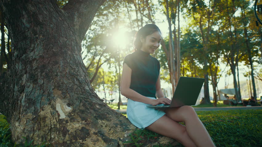 Young woman wearing casual clothing is sitting under a tree and using a laptop. The sun is shining through the leaves, creating a dappled light effect