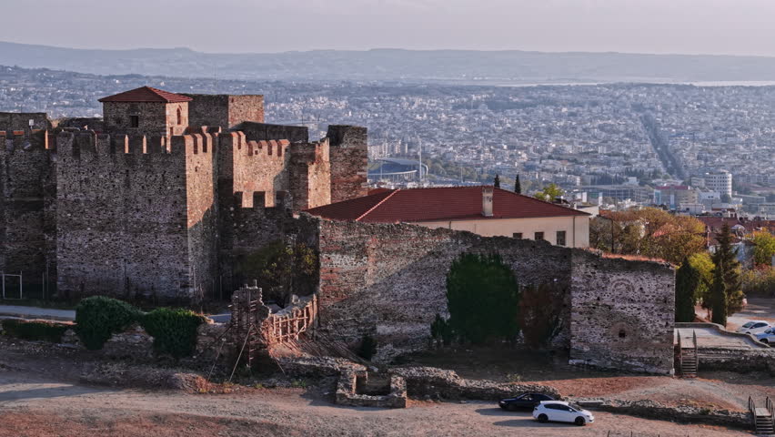 An aerial view captures a historic fortress in Thessaloniki, surrounded by a sprawling urban landscape under a soft evening light, highlighting rich cultural heritage.