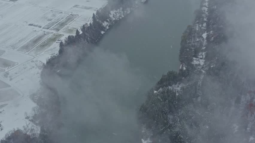 Drone footage: a river flowing at the foot of a snow-covered mountain covered in deep fog
