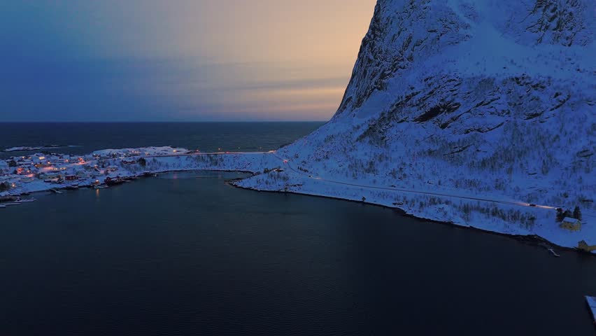 Flying into the sunset at Reine, Lofoten with snow-covered roads and the sun hiding behind the big Reinebringen mountain