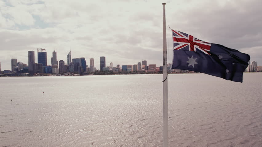 Australian flag waves in the breeze against the Perth skyline and river under gray skies.