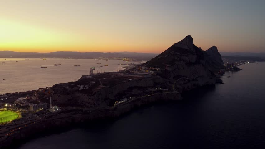Aerial drone view of Gibraltar at sunset. Street lights on. Europa Sports Complex Stadium with night light on. Famous travel destination. Bay of Gibraltar. Orange and pink sunset. drone tracking left.
