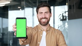 Handsome businessman presenting mobile phone with green screen standing in modern business office. Color key, vertical template layout for advertising. Smiling man looking at camera holding smartphone - Powered by Shutterstock - Get 15% off with code: PIKWIZARD15