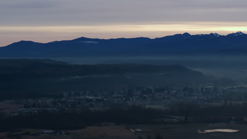 Scenic view of Mount Rainier at sunrise, overlooking a mist-covered valley and distant rural houses, with dramatic lighting and soft atmospheric hues.