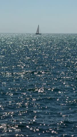 Blue sea sparkles with the reflection of the sun, sailing boat in the ocean, vertical video.