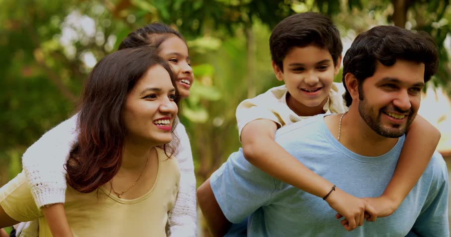 Indian asian Happy Young Family of Four Bonding Outdoors in a Park, Parents Giving Piggyback Rides to Their Smiling Son and Daughter, Walking Amidst Nature and Talking