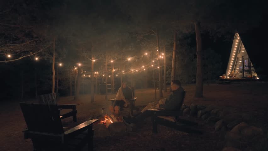 Cozy evening by the fire. A girl and a guy are sitting by the fire. Rest in the forest near a triangular house. A young couple is chatting lively.
