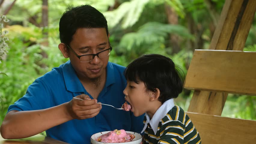 A joyful father and son bonding moment outdoors. The smiling father feeds his child with a fork while sitting together on a wooden bench surrounded by lush greenery,showcasing love and family happines