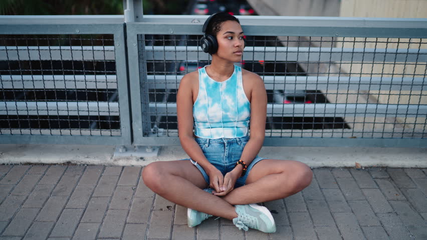 Young depressed woman sitting cross-legged on urban pavement, wearing headphones and casual denim shorts. Relaxed expression, enjoying music in city setting. Modern street style and serene moment