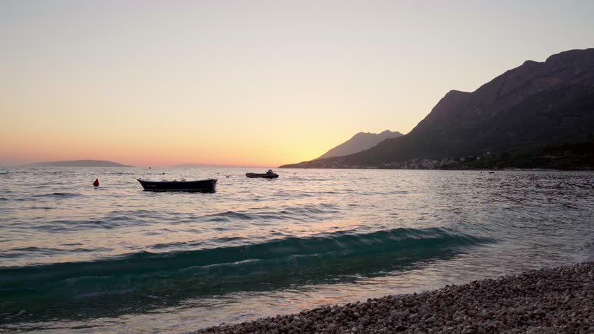 Sunset over the calm sea in Gradac, Croatia. There are two small boats anchored in the water and a mountain range in the distance.