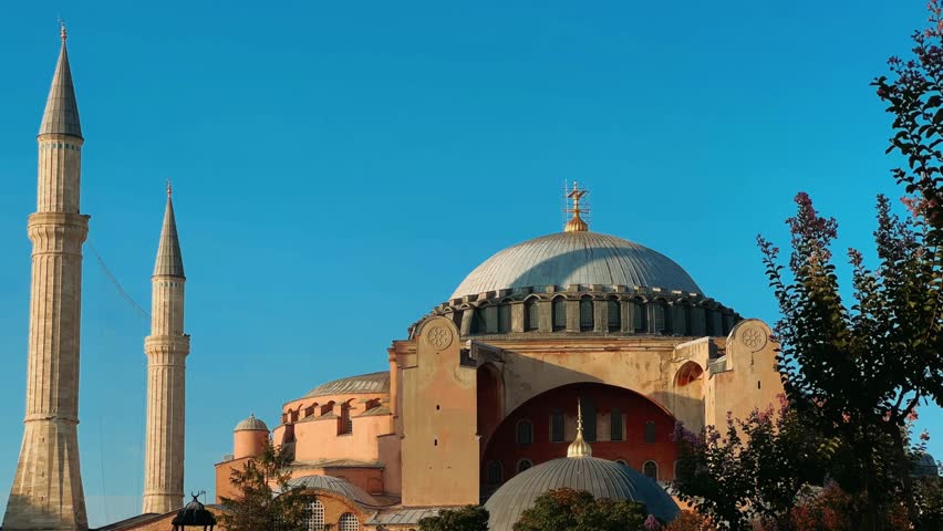 ISTANBUL, TURKEY - JULY 15, 2024: The beautiful Hagia Sophia and the fountain with trees and flower beds in the park. The Christian Patriarchal Basilica, the Imperial Mosque against the blue sky. 4K
