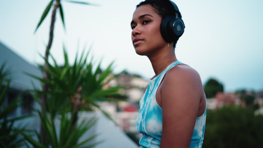 Close-up of young African American woman wearing modern black wireless headphones, gazing upward with thoughtful expression. serene sky creates calming evening background, moment of introspection