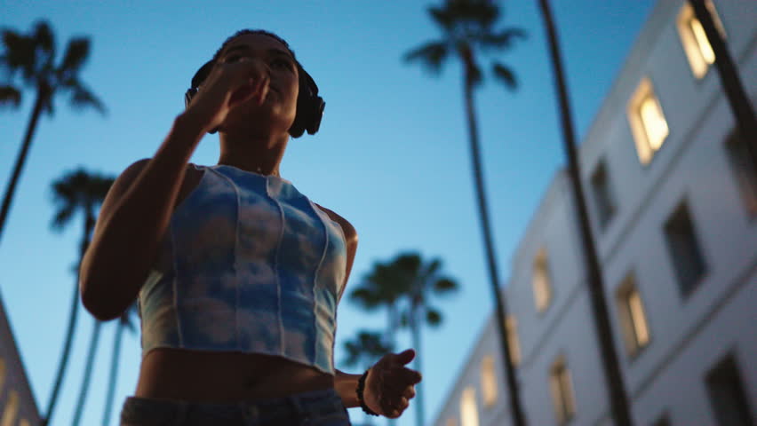 determined young Black woman runs in evening, wearing headphones and listen to music. Focused and motivated, she trains outdoors in a modern urban courtyard with palm trees and glowing windows