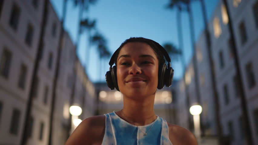 young Black woman with short curly hair enjoys music through wireless headphones while standing in city during evening twilight. African American female listen favorite beat outdoors in earbuds