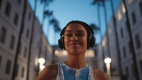 young Black woman with short curly hair enjoys music through wireless headphones while standing in city during evening twilight. African American female listen favorite beat outdoors in earbuds - Powered by Shutterstock - Get 15% off with code: PIKWIZARD15
