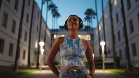Relaxed young dark skinned woman listens to music through stylish black headphones, standing in modern urban courtyard during twilight. eyes are closed, she smiles peacefully, fully immersed in music - Powered by Shutterstock - Get 15% off with code: PIKWIZARD15