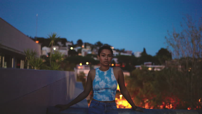 Beautiful young woman stands on twilight rooftop, soft city lights flickering behind her. Her calm gaze meets lens, embodying quiet strength and introspection. sky mirrors her tranquil, untold story